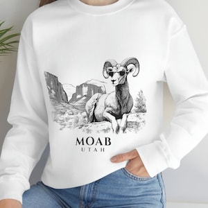 May include: White sweatshirt featuring a black and white illustration of a bighorn sheep wearing sunglasses, with the text "MOAB UTAH" below. The graphic is set against a desert landscape. The sweatshirt has long sleeves.
