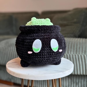 Puede incluir: Un caldero negro tejido a ganchillo con ojos blancos y verdes, mejillas rosas y una sustancia verde claro que se desborda de la parte superior. El caldero tiene tres patas pequeñas y está sobre una mesa blanca.