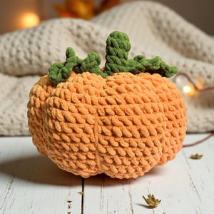 May include: A plush, orange crochet pumpkin with green stem and leaves. The pumpkin is made of a soft, textured yarn and sits on a white wooden surface. The background includes a cozy blanket and soft lighting.