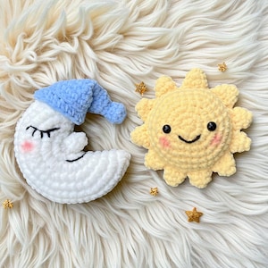 May include: Crocheted moon and sun plush toys. The white moon wears a blue sleep cap and has closed eyes and rosy cheeks. The yellow sun has a smiling face and rosy cheeks. Small gold star-shaped embellishments are scattered around the toys.