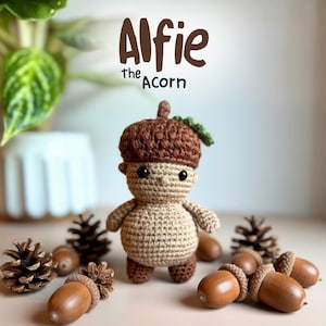 Puede incluir: Muñeca de ganchillo con forma de bellota, con gorro marrón y cuerpo beige. La muñeca tiene brazos y piernas pequeños, y ojos negros. El texto "Alfie the Acorn" está encima de la muñeca. Bellotas y piñas rodean la muñeca.