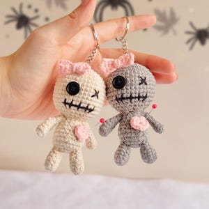 May include: Two handmade crochet voodoo doll keyrings. One is off-white, the other grey, each with a pink bow, black button eyes, and a stitched mouth. A pink heart is pinned to each doll.