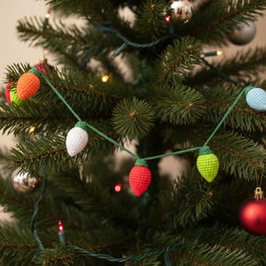 Op de afbeelding: Een handgemaakte gehaakte kerstlichtslinger met kleurrijke lampen. De slinger heeft oranje, witte, rode, groene en blauwe lampen, elk met een groene basis, geregen aan een groen koord. De slinger wordt getoond op een versierde kerstboom.