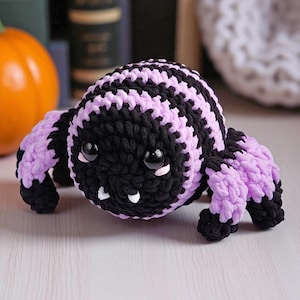 May include: A handmade crochet spider in black and lavender. The spider has large, black eyes, small white fangs, and lavender legs. The spider is a Halloween or fall-themed decoration.