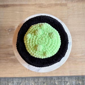 May include: A handmade crochet item featuring a black outer ring and a light green center with raised, textured details. The item is circular and sits on a white and gray marble-like surface.
