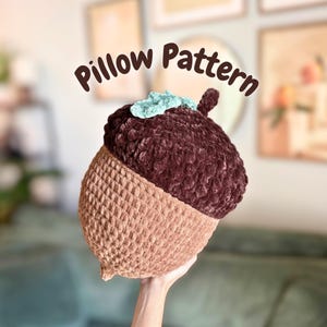 May include: A handmade acorn-shaped pillow in shades of brown and tan. The top of the pillow is a deep brown, plush material, while the bottom is a lighter tan, textured fabric. The pillow has a small, light blue accent and the text "Pillow Pattern".