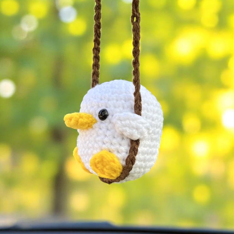 K&ouml;nnte beinhalten: Ein handgeh&auml;keltes, wei&szlig;es Vogel-Ornament mit gelbem Schnabel und F&uuml;&szlig;en, das an einer braunen H&auml;kelschnur h&auml;ngt. Der Vogel hat ein schwarzes Auge und ist vor einem verschwommenen gr&uuml;n-gelben Hintergrund platziert.