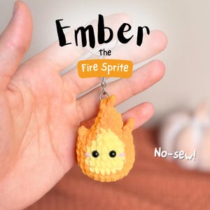 May include: A handmade, no-sew, crochet keychain depicting a fire sprite named Ember. The keychain is predominantly orange and yellow, with black eyes and pink cheeks. The text "Ember the Fire Sprite" is above the keychain.