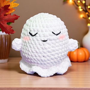 May include: A handmade white crochet ghost with black eyes and a smiling mouth. The ghost has rosy cheeks and small arms. A small orange pumpkin sits nearby, suggesting a Halloween theme.