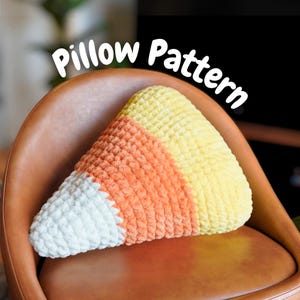 Puede incluir: Una almohada en forma de caramelo de maíz en blanco, naranja y amarillo, apoyada en una silla marrón. La almohada tiene un aspecto texturizado y afelpado. Las palabras "Pillow Pattern" están arqueadas sobre la almohada en texto blanco.