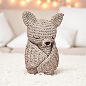 May include: A crocheted plush bat toy with closed eyes and a brown nose. The bat is sitting on a white fluffy surface.