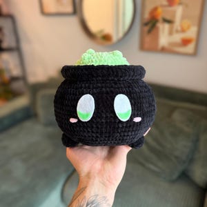 May include: A black crocheted cauldron with white and green eyes and pink cheeks, holding a light green crocheted frog. The cauldron has a round shape and small feet. The plush toy is held in a hand.