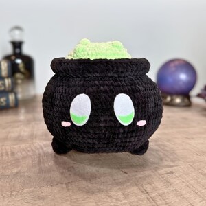 May include: A black crochet cauldron with white and green eyes, pink cheeks, and a light green, textured filling. The handmade plush toy has a whimsical design, perfect for Halloween or fantasy-themed decor.