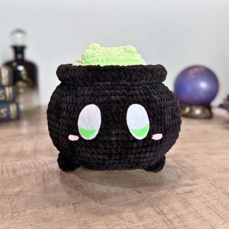 May include: A black crochet cauldron with white and green eyes, pink cheeks, and a light green, textured filling. The handmade plush toy has a whimsical design, perfect for Halloween or fantasy-themed decor.