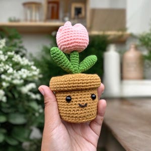 Peut inclure: Un pot de fleurs en crochet avec une tulipe rose et un visage souriant. Le pot est brun et les feuilles sont vertes.