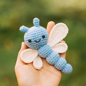 Può includere: Un giocattolo a forma di farfalla all'uncinetto in azzurro e crema. La farfalla ha un viso sorridente con occhi neri e un corpo segmentato. Le ali sono color crema e il corpo azzurro. Il giocattolo è tenuto in mano su uno sfondo verde sfocato.