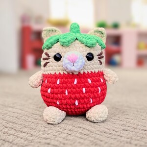 May include: A crocheted stuffed toy shaped like a cat wearing a strawberry costume. The cat has a green crocheted leaf on its head and a pink nose.