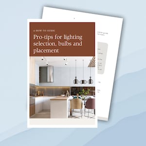May include: A how-to guide on lighting selection, bulbs, and placement for home decor. The image shows a modern kitchen with white cabinets, a stainless steel range hood, and a wooden kitchen island with bar stools. There are three black pendant lights hanging over the island.
