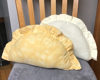 May include: Two plush, dumpling-shaped pillows in the image. One is a light yellow with a mottled pattern, and the other is a solid off-white. They rest on a gray upholstered chair, suggesting a home decor theme.