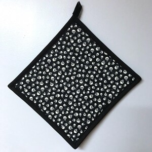 May include: Black and white pot holder with a skull pattern. The pot holder has a black border and a loop for hanging.
