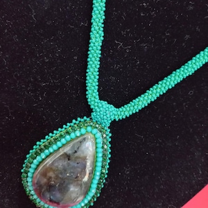 May include: A teal beaded necklace with a teardrop-shaped pendant featuring a large, iridescent labradorite stone surrounded by smaller teal beads.