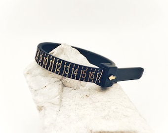 Pode incluir: Pulseira de fita métrica preta e dourada com os números de 8 a 17. A pulseira é feita de plástico flexível e tem um fecho de fivela.