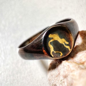 May include: A silver signet ring with a black oval top featuring a gold silhouette of a person. The ring is resting on a light brown rock.