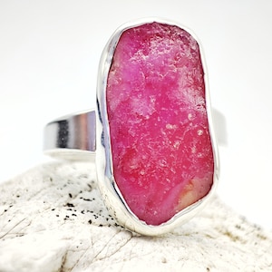 Peut inclure: Une bague en argent avec une grande pierre précieuse de rubis brut de couleur rose sertie au centre. La bague est posée sur une surface blanche texturée.