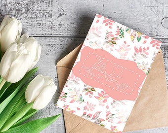 Peut inclure: Une carte rose et blanche avec un motif floral et le texte "The best Mom" dans un cadre blanc en forme de cœur. La carte est posée sur une enveloppe brune avec un bouquet de tulipes blanches en arrière-plan.