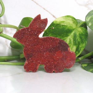 May include: A red, glitter-covered bunny-shaped decoration. The bunny is a solid color with a textured surface. The background includes green leafy plants and a white marble surface.