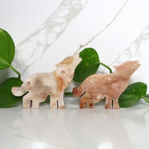 Puede incluir: Dos figuritas de lobo talladas en cristal, una aullando y la otra con la cabeza girada hacia un lado. Los lobos están hechos de un cristal marrón claro y rosa con un acabado brillante.