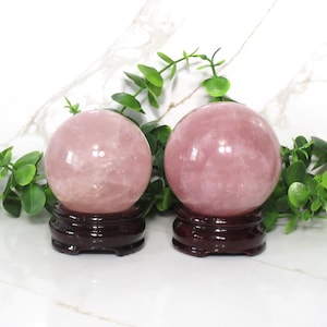 May include: Two pink rose quartz spheres on wooden stands. The spheres are polished and have a translucent appearance.