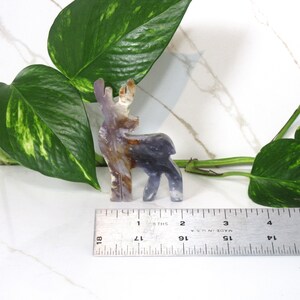 May include: A small, carved deer figurine made of agate. The deer is standing on a white surface with a ruler for scale. The deer is about 2 inches tall.