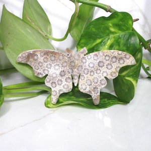 May include: A butterfly-shaped decorative object with a detailed floral pattern in shades of beige and brown. The object is resting on a green leaf, with additional green foliage in the background. The butterfly has a textured appearance.