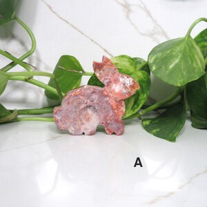 May include: A carved stone bunny figurine in shades of red, pink, and white. The bunny is positioned on a white surface with green foliage in the background. The letter "A" is visible in the lower right corner.