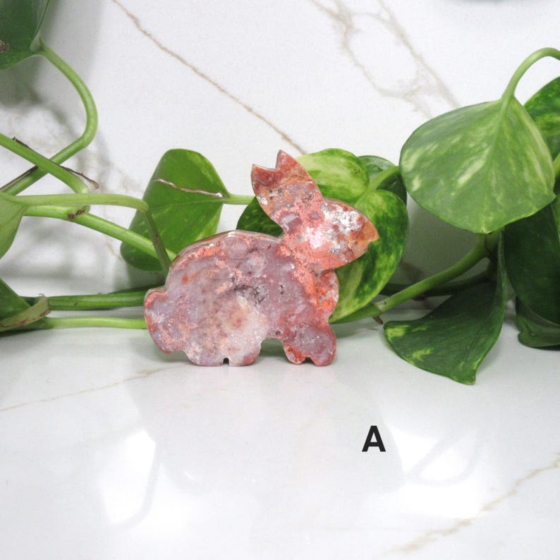 May include: A carved stone bunny figurine in shades of red, pink, and white. The bunny is positioned on a white surface with green foliage in the background. The letter "A" is visible in the lower right corner.