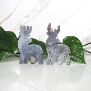 May include: Two gray reindeer figurines made of a marbled material. The reindeer are standing on a white surface with green leaves in the background.