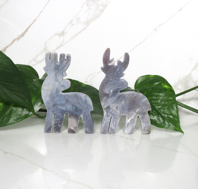 May include: Two gray reindeer figurines made of a marbled material. The reindeer are standing on a white surface with green leaves in the background.