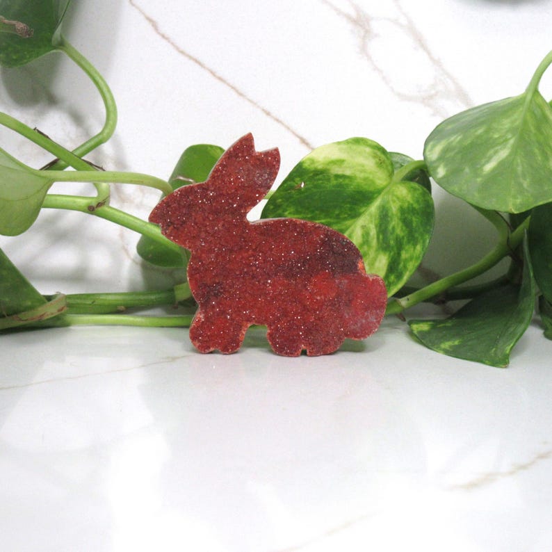 May include: A red, glitter-covered bunny-shaped decoration. The bunny is a solid color with a textured surface. The background includes green leafy plants and a white marble surface.