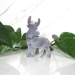 May include: A gray and white marble-like reindeer figurine. The reindeer is standing on a white surface with green leaves in the background.