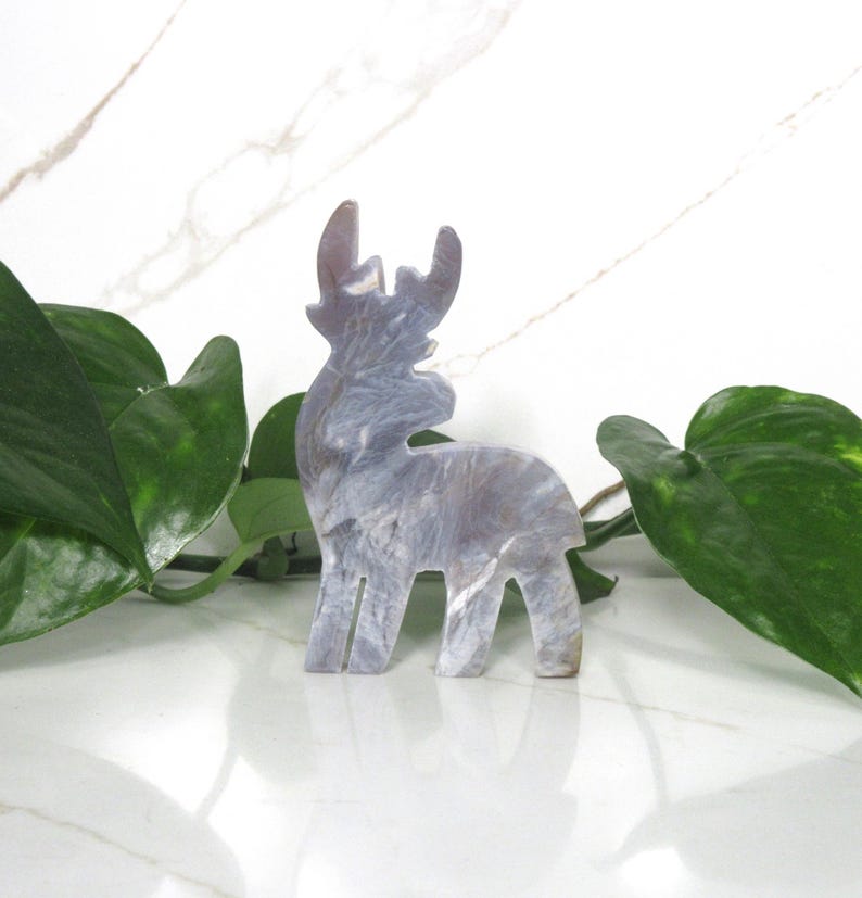 May include: A gray and white marble-like reindeer figurine. The reindeer is standing on a white surface with green leaves in the background.