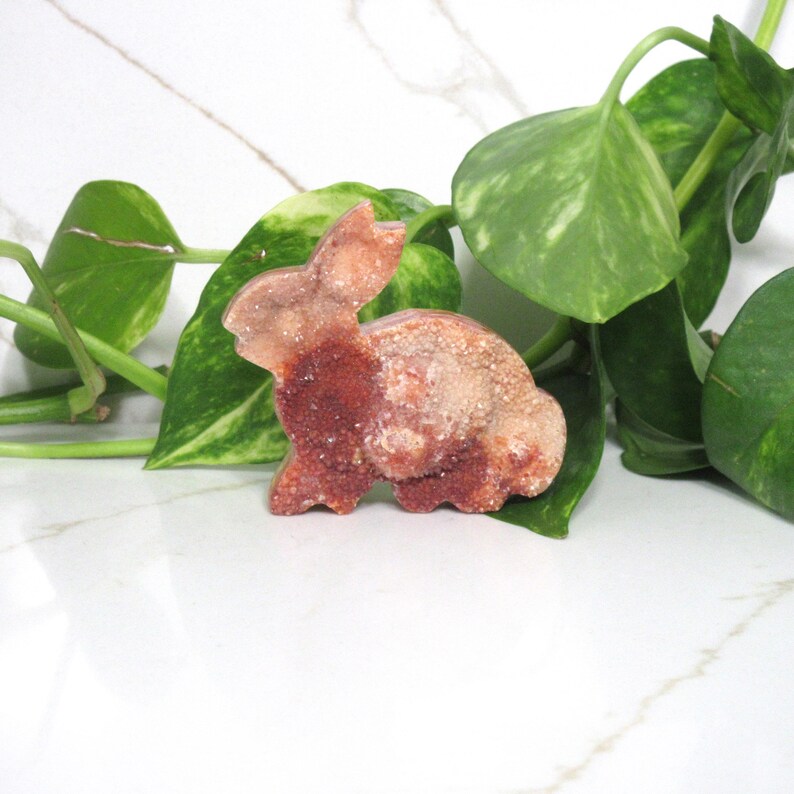May include: A reddish-brown crystal rabbit figurine with a textured surface. The bunny is set against a backdrop of green leaves and a white marble surface. The figurine is a decorative item.
