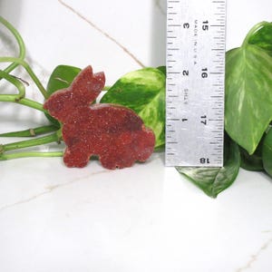 May include: A red-brown crystal bunny figurine, approximately 2 inches in length, sits next to a ruler. The bunny is textured and sparkly, with a detailed outline. Green leaves and a white marble surface provide the background.