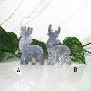 May include: Two gray, marble-like reindeer figurines. The reindeer are standing on a white surface with green leaves behind them. The reindeer are labeled A and B.