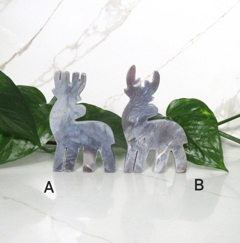 May include: Two gray, marble-like reindeer figurines. The reindeer are standing on a white surface with green leaves behind them. The reindeer are labeled A and B.
