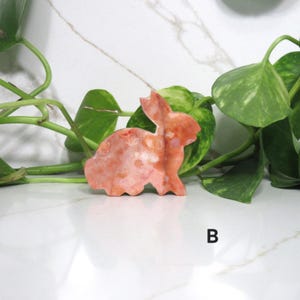 May include: A carved stone bunny figurine in shades of orange and pink. The bunny is posed in a sitting position with its ears perked up. The figurine is set against a backdrop of green leaves and a white marble surface.