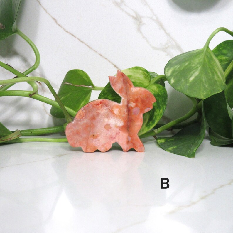 May include: A carved stone bunny figurine in shades of orange and pink. The bunny is posed in a sitting position with its ears perked up. The figurine is set against a backdrop of green leaves and a white marble surface.