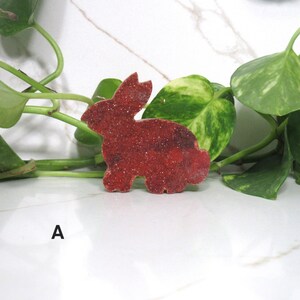 May include: A red, glitter-covered bunny-shaped decoration. The bunny is positioned on a white surface, with green leafy vines in the background. The letter "A" is printed in black on the lower left.