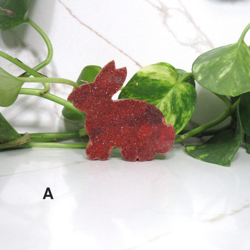 May include: A red, glitter-covered bunny-shaped decoration. The bunny is positioned on a white surface, with green leafy vines in the background. The letter "A" is printed in black on the lower left.