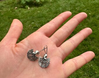May include: A pair of silver-colored dangle earrings. Each earring features a detailed, textured design resembling a flower or a stylized burst. The earrings are displayed on an open palm against a backdrop of green grass.